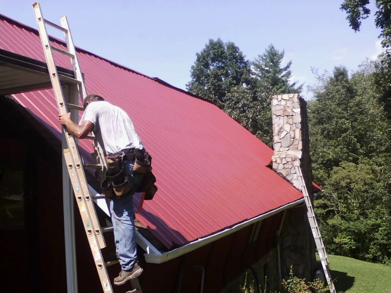 Roofer on a ladder beside a red metal roof and stone chimney — Soffit & Fascia Repair by Roofing Brothers in Neah Bay